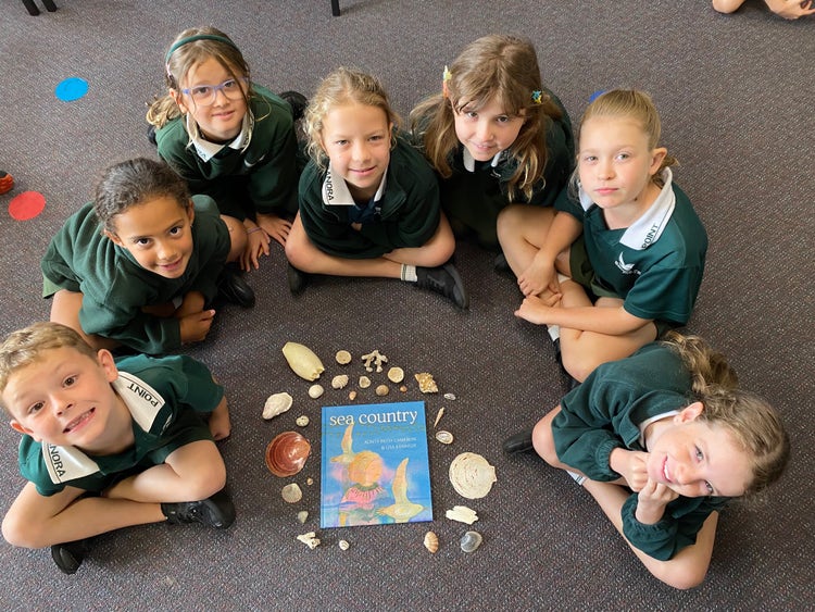 A small group of students sitting around their class text 'Sea Country.' There is a collection of shells surrounding the text which the students collected from the beach.